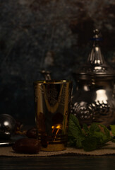 vertical photo of a glass with arabic motifs filled with tea and illuminated from the inside and silver plates with brown sugar or mint with dates and tea on one of the plates dark photo  