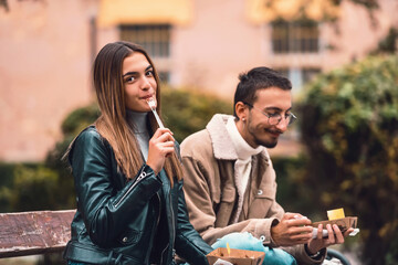Two students modernly dressed sit in the park on a break from college and eat candy. Selective focus