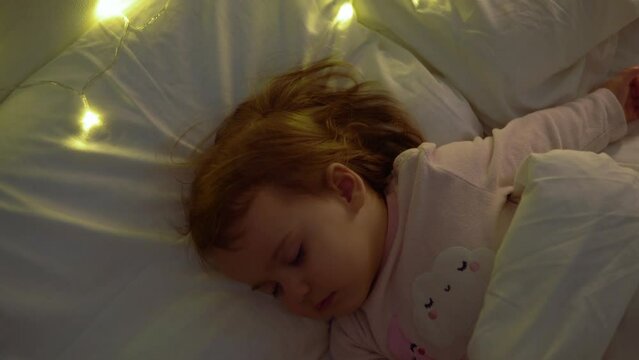 Cute Chuby Little Toddler Baby Girl 1-2 Years Old Sleeping In Pink Pajama On White Bed In Dark Bedroom. Child Have Relax Rest Time At Night Garland on Pillow. Childhood, Christmas, Parenthhood Concept