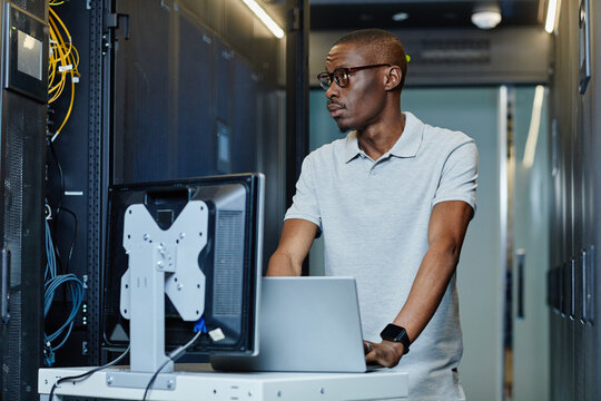 Portrait of African American man connecting laptop to server in server room and setting up internet network, copy space