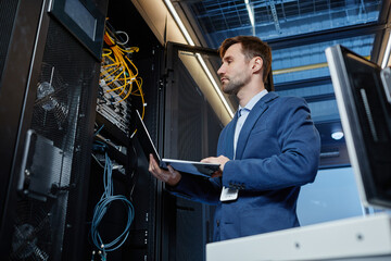 Low angle portrait of IT engineer holding laptop while setting up internet network in server room