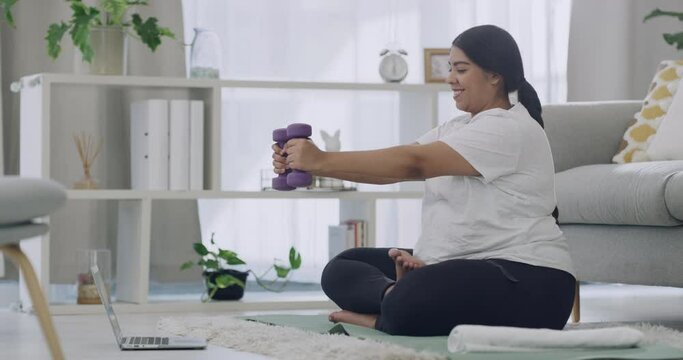 One happy mixed race plus size female model starting weight loss journey in an apartment on the lounge floor. Latino woman watching a fitness tutorial on a laptop while exercising at home