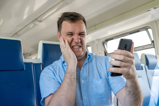 Sad millennial guy looks at phone screen in surprise, sits in commuter train. Disgusted and overwhelmed man stares at screen of smartphone with confuse, bad joke or inappropriate content