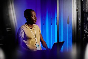 Side view portrait of adult black man using laptop in dark server room lit by neon light, copy space