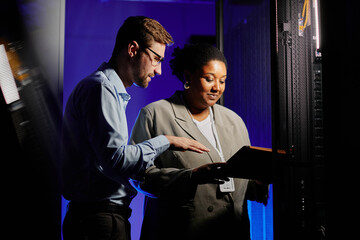 Waist up portrait of two system administrators using computer in dark server room lit by neon...