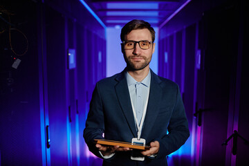 Waist up portrait of male network engineer holding tablet in server room and looking at camera