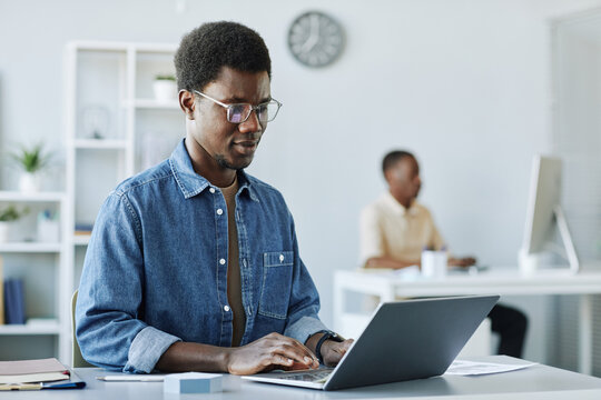 Portrait of young African American man working in office and using laptop in minimal grey interior, copy space