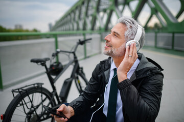 Businessman with bike sitting on bench, listening to music and resting. Commuting and alternative transport concept