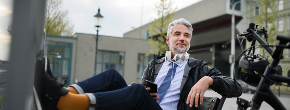 Businessman with bike sitting on bench, listening to music with feet up and resting. Commuting and alternative transport concept
