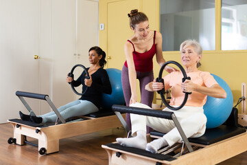 Fototapeta na wymiar Young female pilates instructor assisting elderly woman to do exercises on reformer with flex ring and fitball. Active lifestyle concept