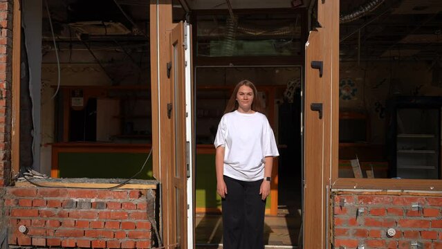 Young woman looking at camera standing at ruined building outdoors. Live camera zoom out to debris in bombed destroyed city in Ukraine. War consequences and military destruction