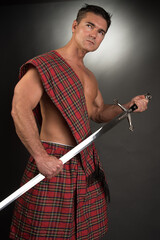 The sexy highlander man poses in a studio with a dark background setting. He holds a sword and...