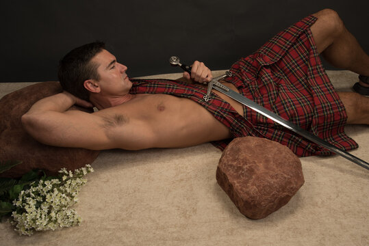 The sexy highlander man poses in a studio with a dark background setting. He holds a sword and prepares himself for the epic battle.