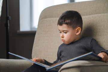 Little boy reading a book while sitting on sofa, at home. Education and early development of children.