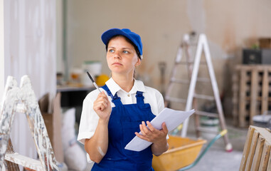 Young woman builder checking project documentation while working in apartment. Girl in coverall...