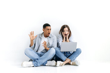 Amazed stunned young multiracial couple, in casual wear, sitting on floor over isolated white background with laptop, looking at a screen in surprise, saw unexpected news, big discount in online store