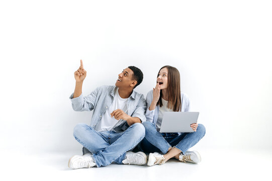 Happy multiracial couple, a hispanic man and a caucasian woman, with a laptop, are looking to the side, the guy points with hand at something, while sit on the floor on white isolated background,smile