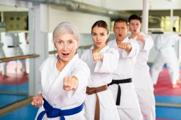 Tuinposter Focused elderly woman wearing kimono standing in attacking stance, practicing movements of close combat punches in training room during group martial arts workout.. © JackF