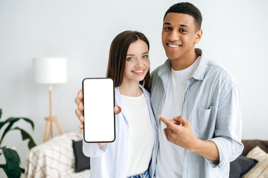 Positive modern caucasian girl and hispanic guy, a couple in love, in stylish clothes, stand in a cozy living room, show a smartphone with a blank white mockup screen, empty space for advertisement - Powered by Adobe