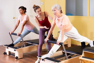 Fototapeta na wymiar Friendly young girl professional pilates instructor assisting elderly woman to do exercises on reformer. Active lifestyle concept