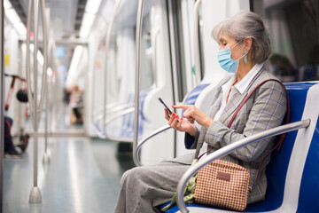 Caucasian mature woman in face mask sitting inside subway train and using her smartphone.