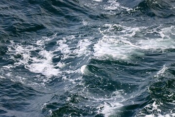 Stern waves with white foam tips on greyish blue sea water, photo taken from aboard ship. Selective focus