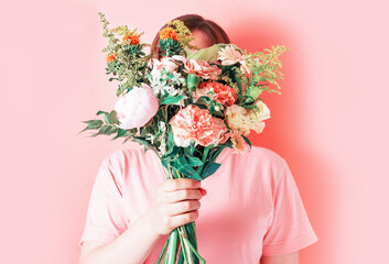 Young woman holding a bouquet of flowers covering her face on a pink background.
