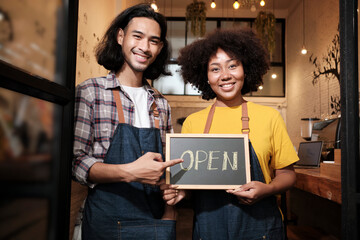  Two young startup barista partners with aprons stand at casual cafe door, letters on board and show open sign, happy and cheerful smiles with coffee shop service jobs, and new business entrepreneurs.