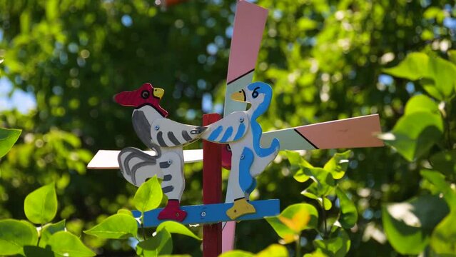A war between the red rooster and the blue, yellow, and white ducks on the pinwheel. Colorful bird windmill toy decoration in the green outdoor garden forest at home during the hot summer heatwave.