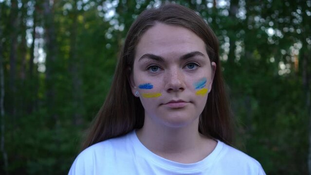 Headshot of young brunette Ukrainian woman with blue and yellow strips painted on face looking at camera. Close-up portrait of sad devastated lady posing outdoors in forest