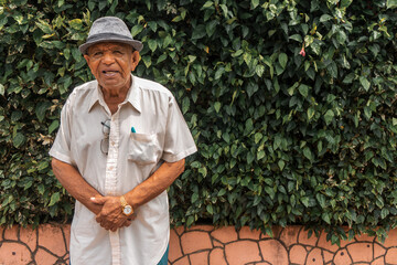 Nicaraguan old man in a hat and short-sleeved shirt looking at the camera with plants in the background