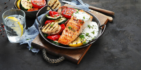 Salmon steak with vegetables and white sauce on dark background.