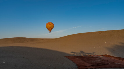 A yellow-red balloon is flying in a clear blue sky over the desert. A carpet is spread out on the sand. light and shadows. Egypt. Luxor
