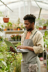 African young gardener in uniform using laptop to look up about plant species during his work in...
