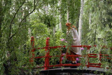 Obraz na płótnie Canvas Happy ginger hair woman waling in well-groomed park or garden with fruits. Freedom and healthy way of life. Female in her 40s having rest outdoors. Cheerfully person on small red bridge . Copy space