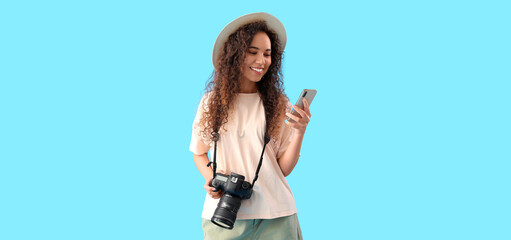 Young female African-American tourist with photo camera and mobile phone on blue background
