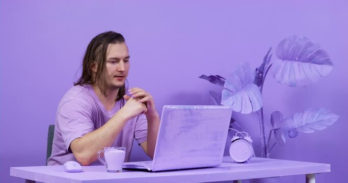 Young man with long hair sighs heavily, getting ready for work, then resolutely opens laptop, turns it on and takes mug from table to inhale pleasant aroma of drink. Everything is painted purple