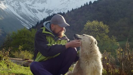Bearded man traveler resting with his dog among beautiful landscape on mountains background. Man stroking his dog and pet sitting next to his owner on glade in forest. Man and dog are best friends