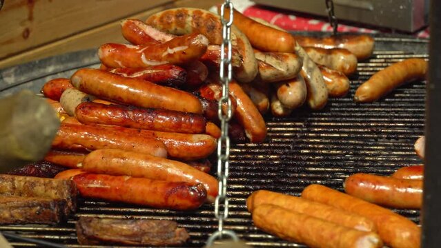 Chef flips sausages Bratwurst roasting with cooking tongs. BBQ grilling frankfurt beef sausage. Street food for picnic and camping. Grilled weisswurst fried and smoked in charcoal grills. 4k footage
