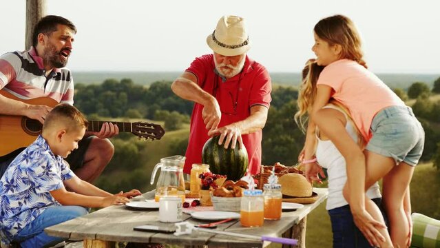 Cheerful Multi Generational Family At Summer Picnic At Table Enjoys Waiting For Grandfather To Cut Watermelon Outdoors In Nature. 