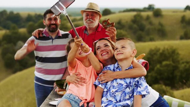 Mother and two small children take a selfie picture with the family using a selfie stick in nature at a picnic in a beautiful hilly landscape