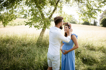 young couple in love standing in high flower meadow in summer and cuddling and the woman is pregnant and has a blue dress on and the man has a full beard