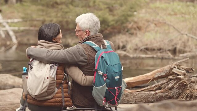 Rear view slowmo of mature active couple with hiking backpacks sitting by lake in forest embracing each other and enjoying nature