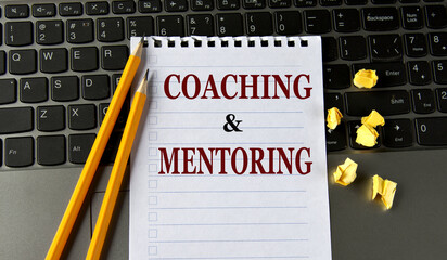 COACHING and MENTORING - words in a white notepad on the background of a laptop with pencils