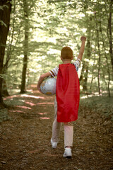 Rear view of caucasian boy wearing superhero costume walking with globe in the forest