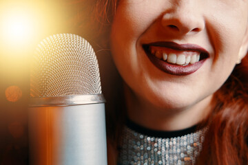 Red-haired cheerful woman with burgundy lipstick on her lips perform at concert. Close-up of smiling female singer's and retro microphone on bokeh blur background. Night party in club or restaurant.