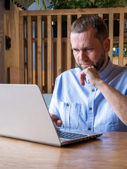 Serious handsome arabian man with beard is using laptop, reading email, typing on keyboard in cafe. Handsome male freelancer working on laptop at workplace. Businessman typing information