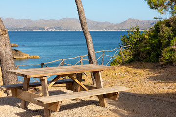 Banco de madera merendero al borde de la playa de s'Illot, en Alcúdia (Islas Baleares, España), con las montañas de la Serra de Tramuntana al fondo.
