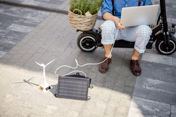 Stylish man works on laptop computer and charges it with solar panels while sitting on electric scooter outdoors. Concept of sustainable lifestyle and green renewable energy. Cropped view