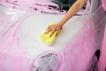 Female person wiping car covered in pink nano foam with yellow sponge at car wash. Close-up view on...